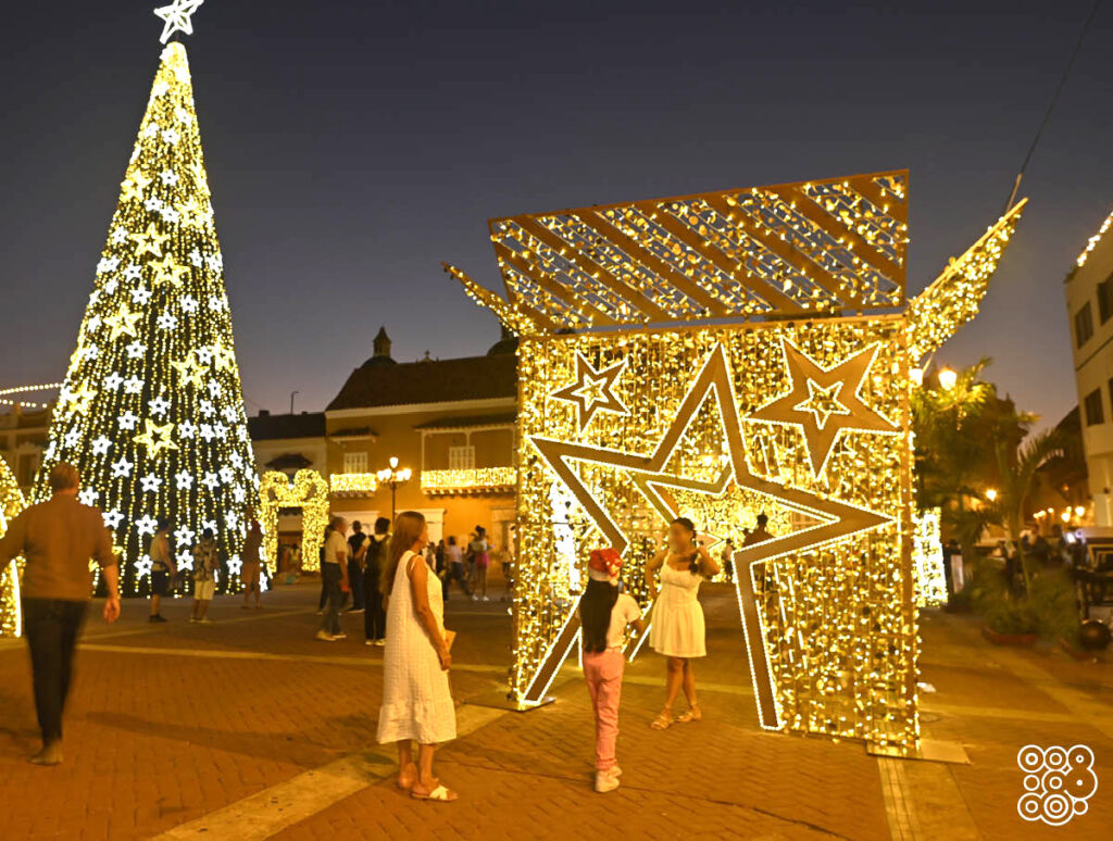 square with people and in the back a giant illuminated christmas tree giant and in the front a walk through illuminated christmas box